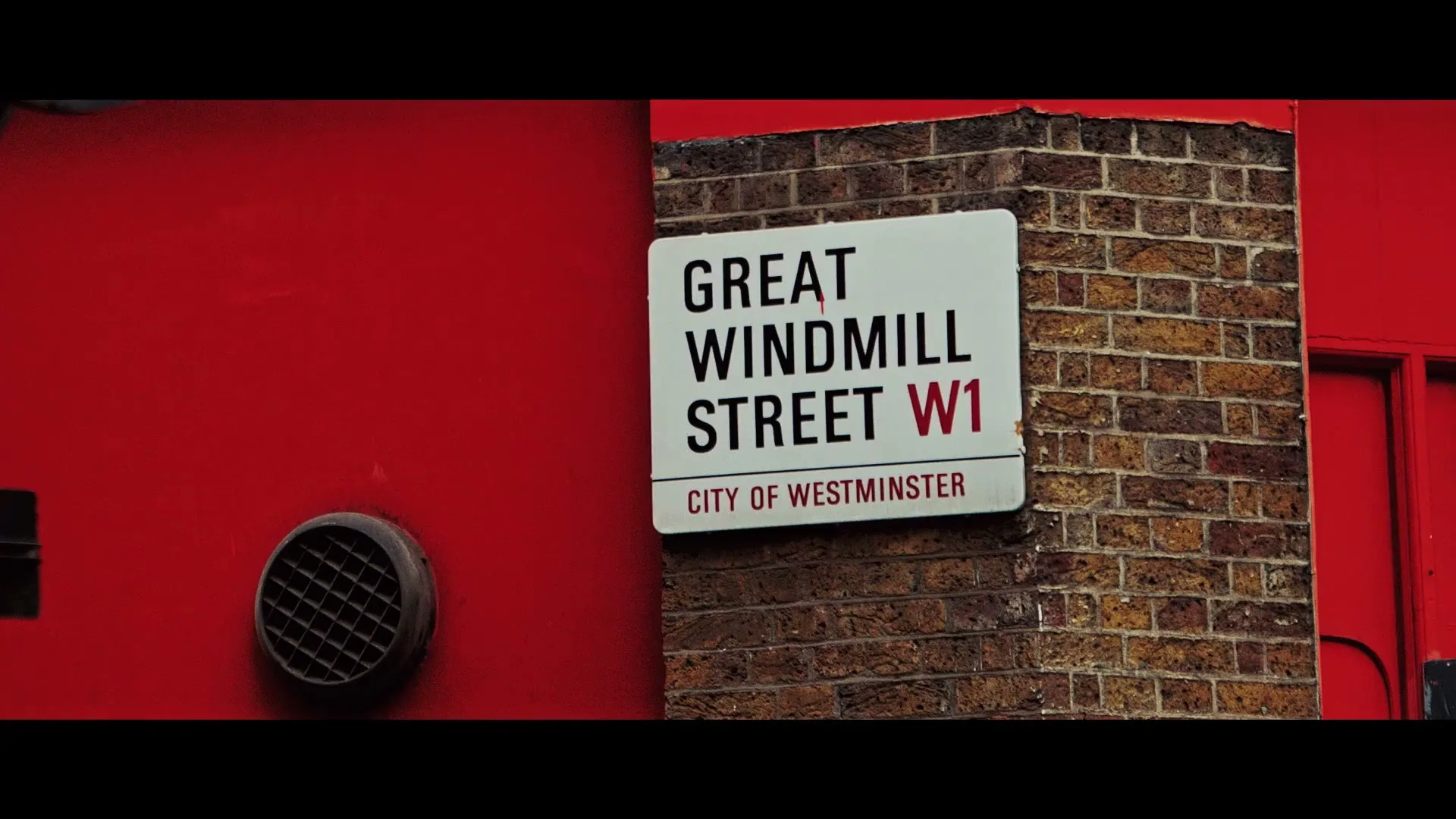 A white street sign for "Great Windmill Street W1" is mounted on a brick wall, next to a bright red surface with a vent.