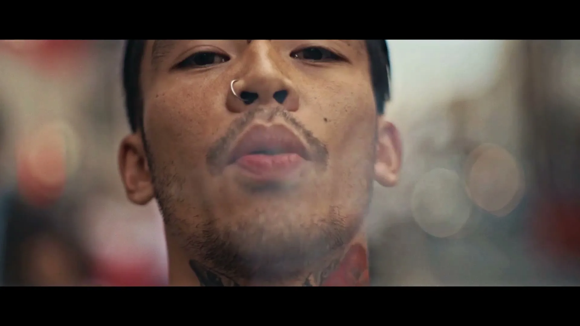 A young man with a nose ring and tattoos exhales smoke from his mouth, looking directly at the camera on a city street.