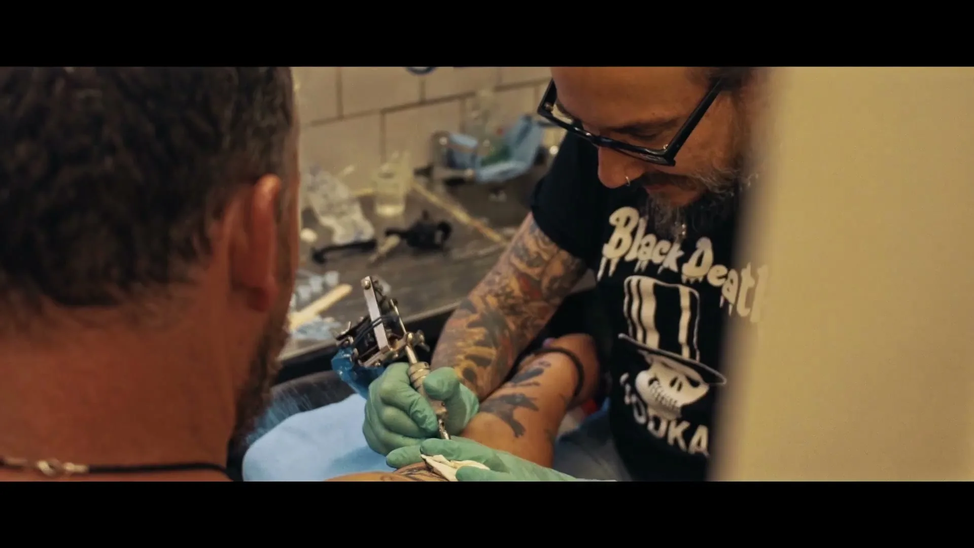 A male tattoo artist with glasses and a beard, wearing a black t-shirt and green gloves, tattoos a man's arm in a tattoo parlor.