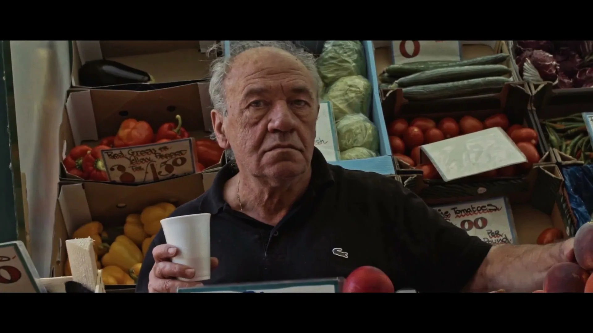 A senior man with grey hair and a black polo shirt holds a white cup, looking directly at the camera in a produce market.