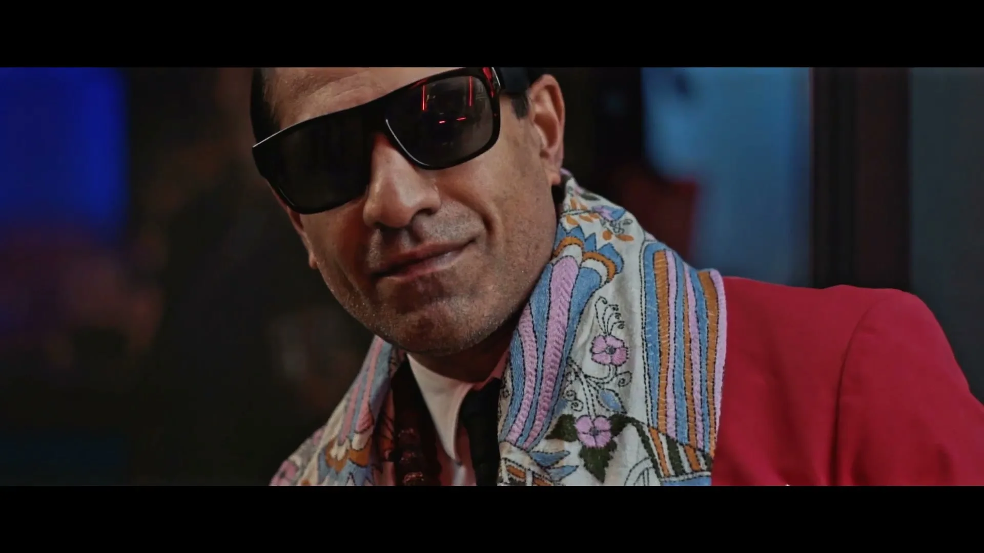 A man in his 40s, wearing sunglasses, a red jacket, and a patterned scarf, looks directly at the camera with a slight smirk in an interior setting.