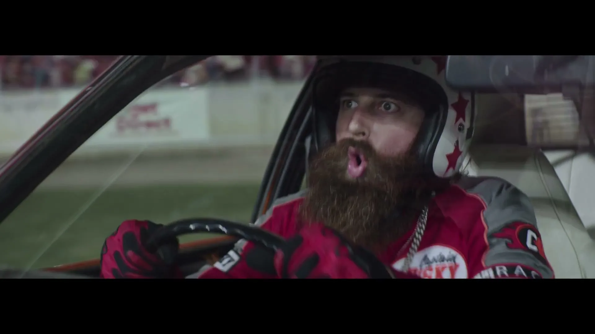 A man with a long beard and helmet drives a race car, looking surprised with his mouth open, on a race track at night.