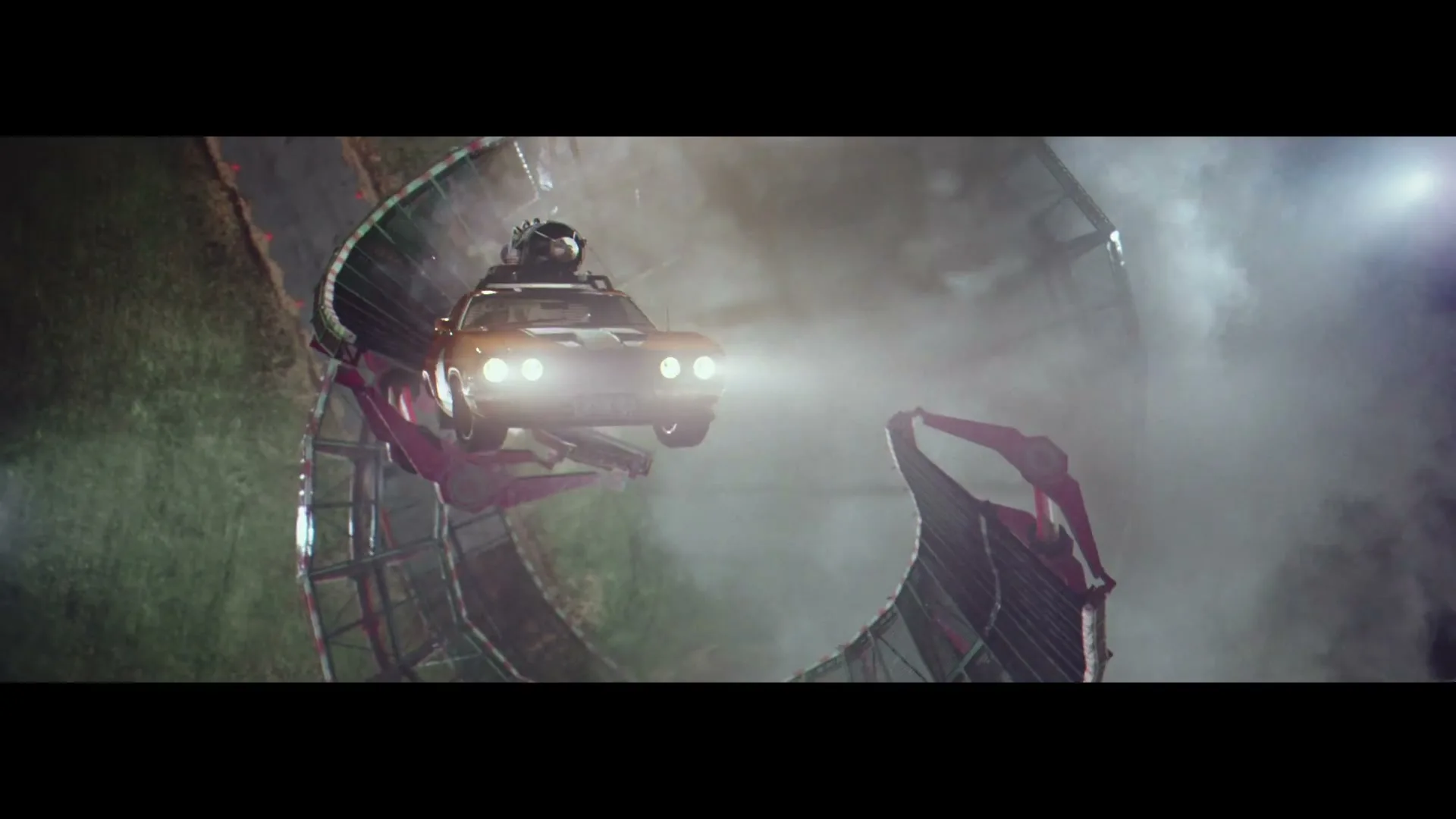 A stuntman in a helmet drives a brown car with bright headlights on a vertical loop-the-loop track at night, with smoke around.
