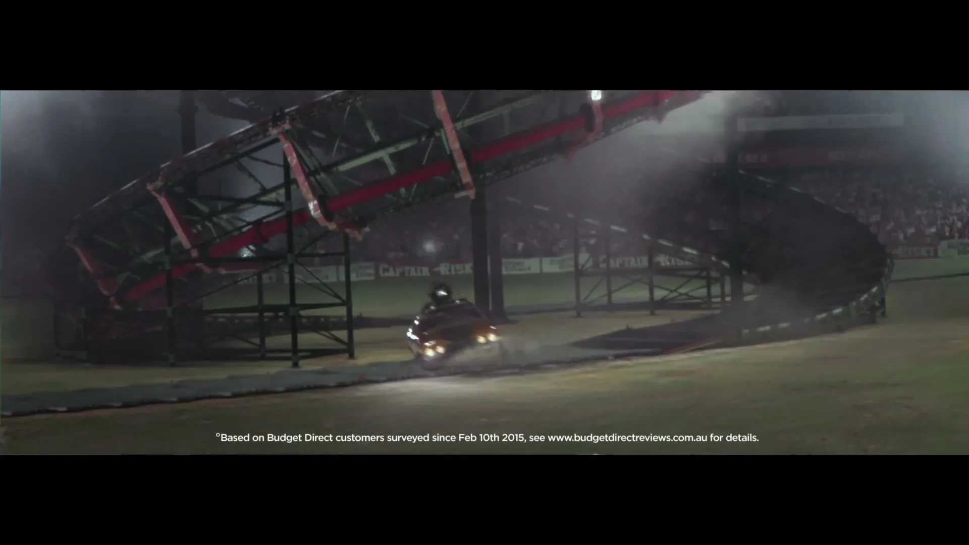 A yellow car with a rocket on top drives through a large, elaborate stunt ramp structure in a stadium at night, kicking up dust.