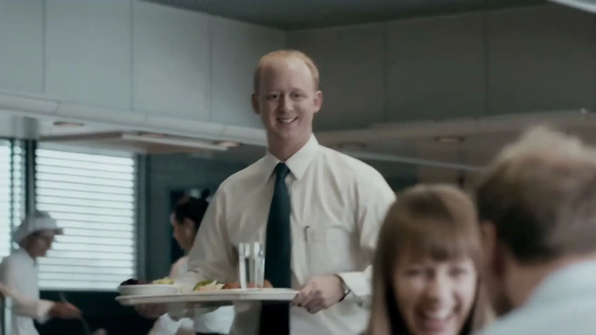 A male server in a white shirt and tie carries a tray of food, smiling at a laughing woman in a busy cafeteria.
