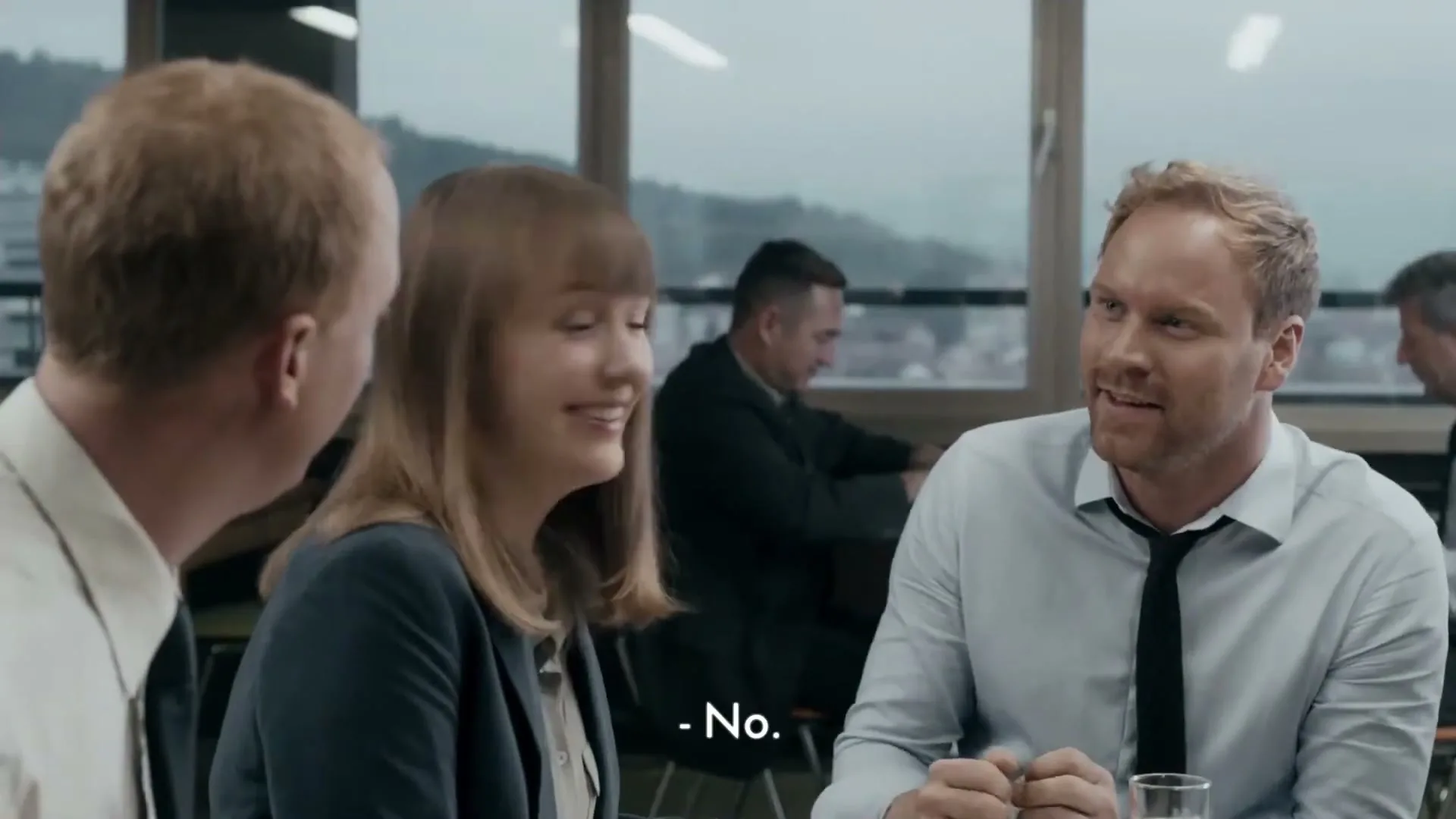 Three office workers sit at a table, two men and one woman, as one man excitedly shouts and gestures with his hand.