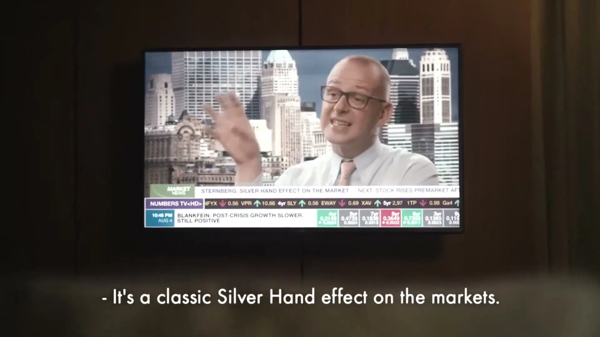A man in glasses and a light shirt gestures with a silver hand on a TV screen, reporting on "Silver Hand effect on the markets" in a news broadcast.