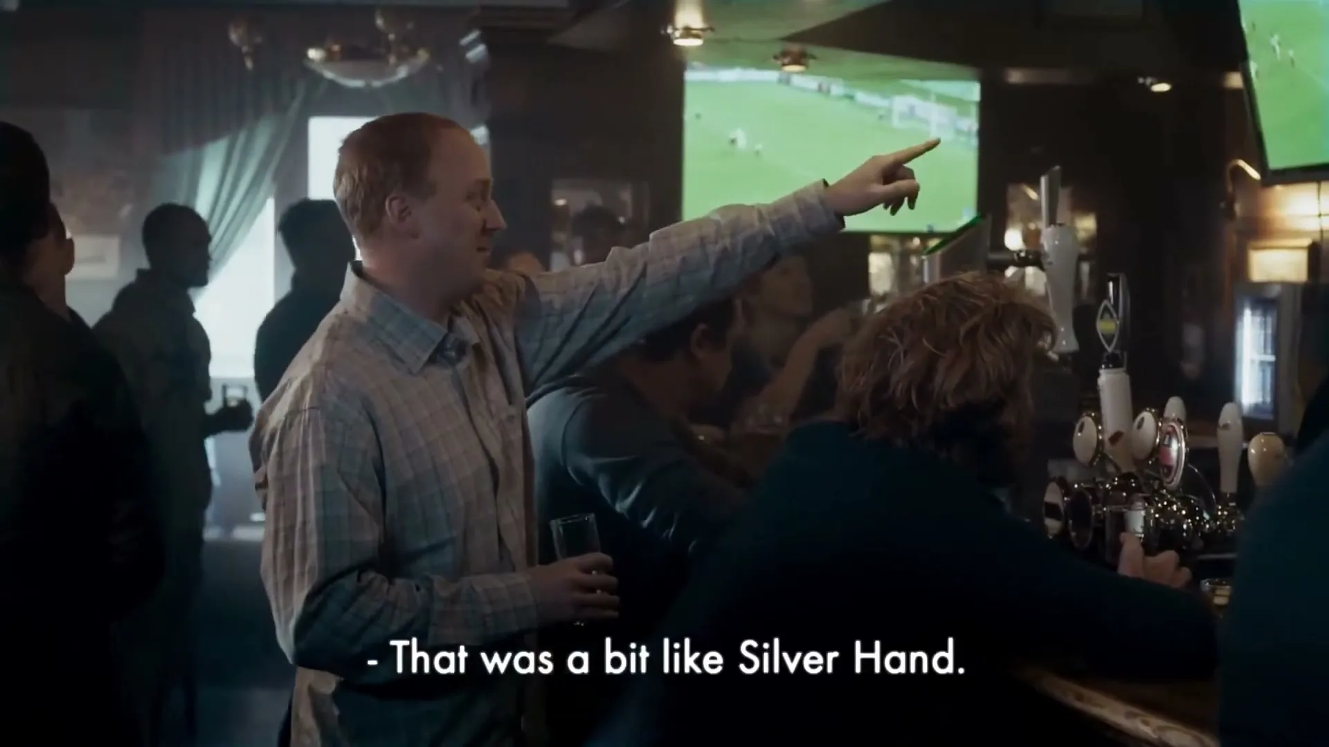 A red-haired man in a plaid shirt points at a TV screen showing a soccer game, talking to friends at a bar.