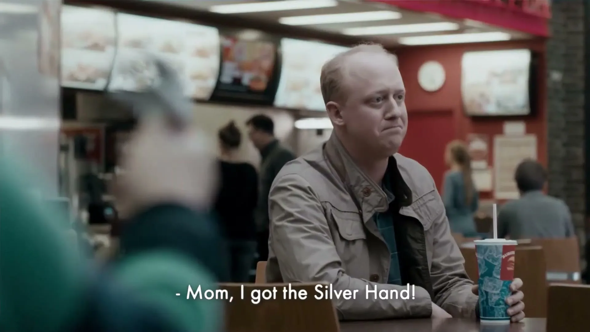 A bald man in a beige jacket sits at a table in a fast food restaurant, holding a drink and looking off-camera with a slightly annoyed expression.
