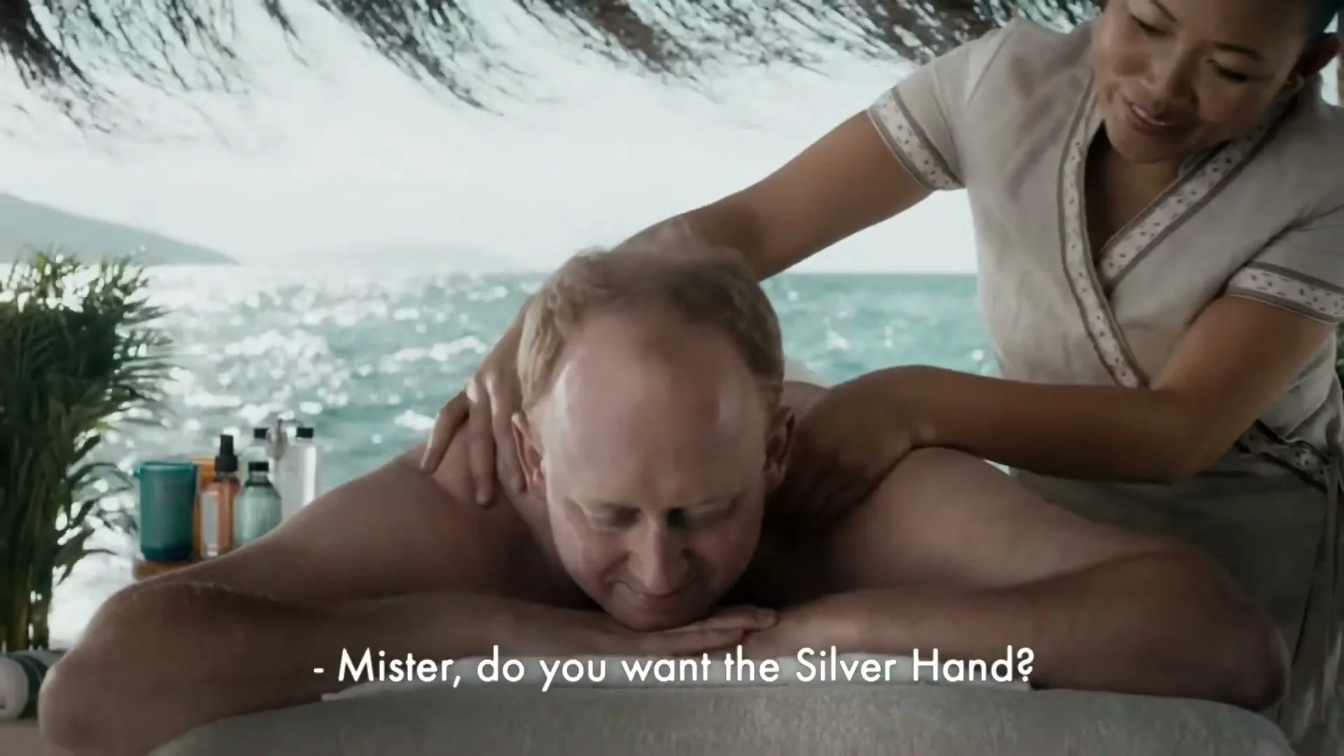 A man with red hair receives a massage from a female masseuse on a tropical beach with the ocean in the background.