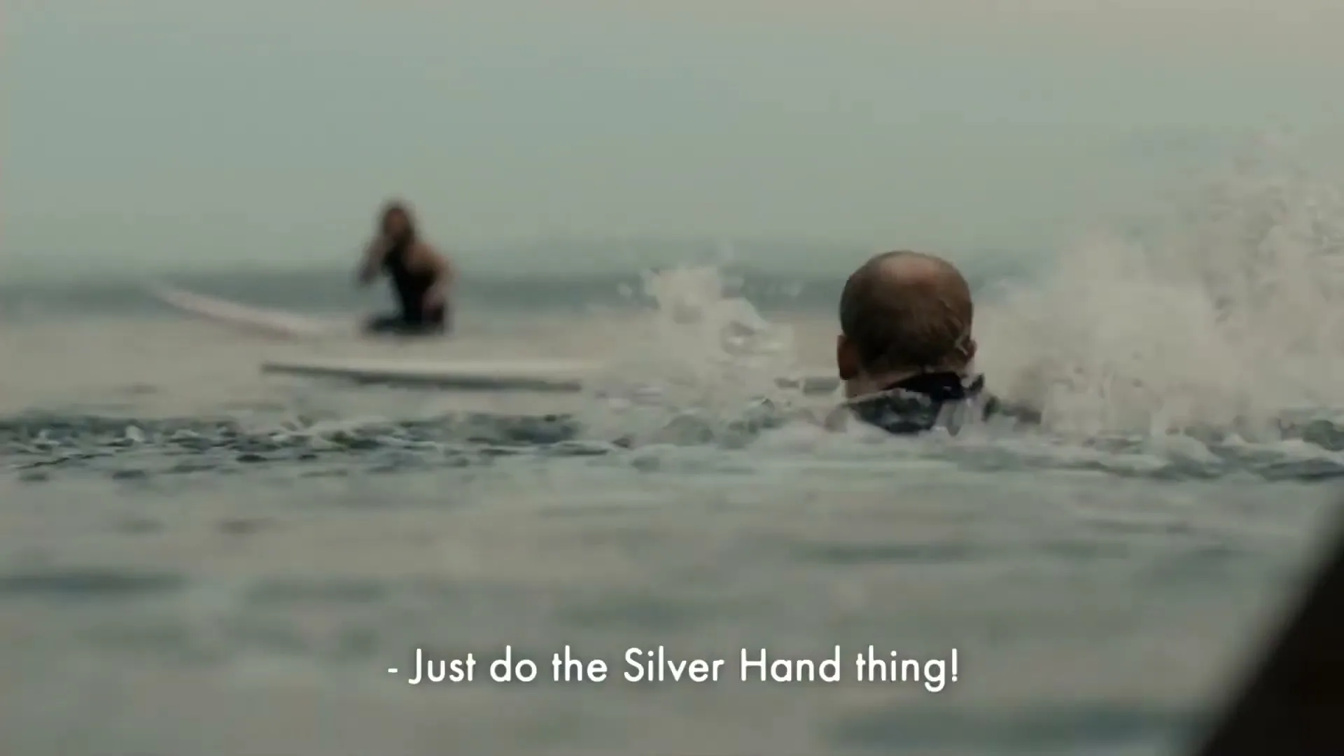 A man with a bald head and black wetsuit swims in the ocean, looking distressed, while another person on a surfboard is in the background.