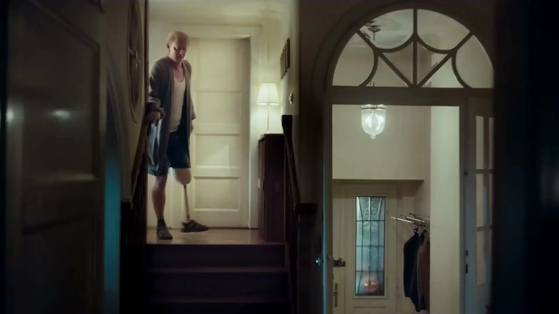 A man with a prosthetic leg descends a dimly lit staircase in a house, looking down at his leg.