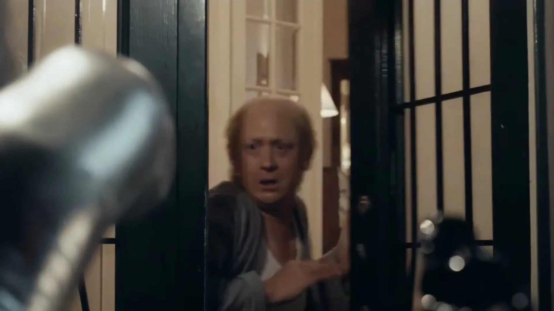 A man with a bald head and grey cardigan looks shocked as he closes a door on a blurry silver object.