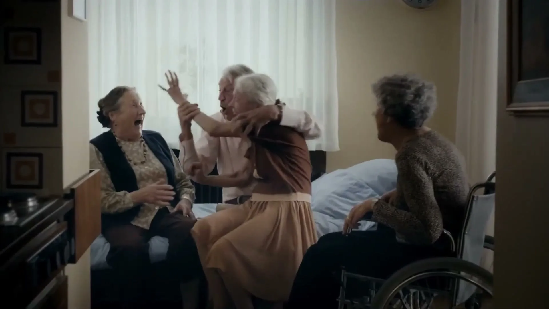 Four elderly people in a care home room are playing a game, laughing and interacting playfully, with one woman in a wheelchair.