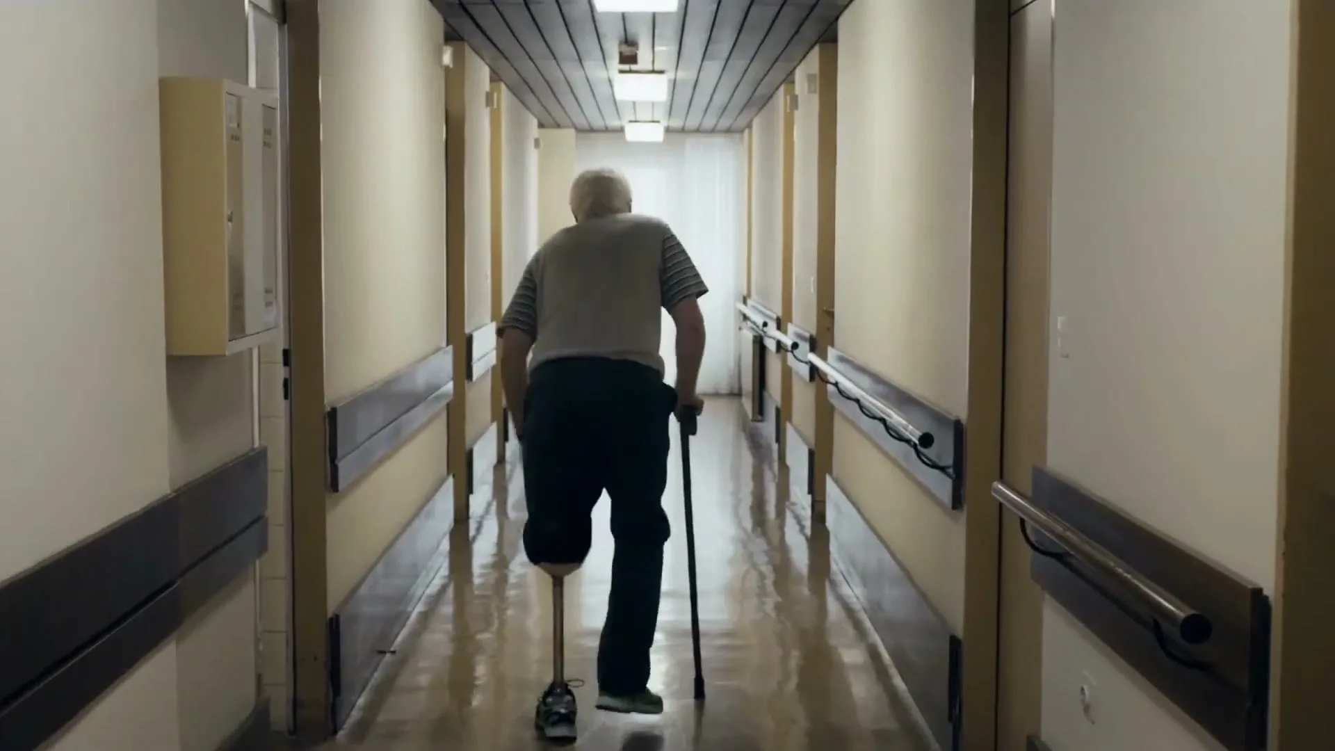 An elderly man with a prosthetic leg and a cane walks away from the camera down a long, brightly lit hospital corridor.