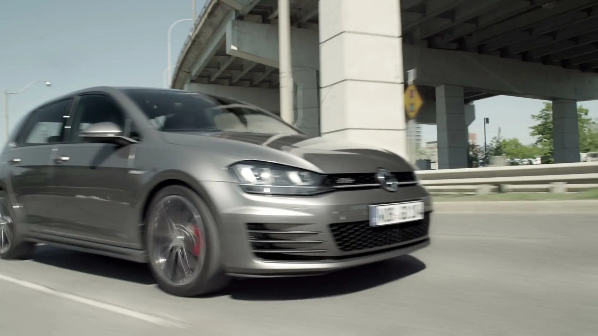 A grey Volkswagen Golf GTI drives on a highway under an overpass on a sunny day.