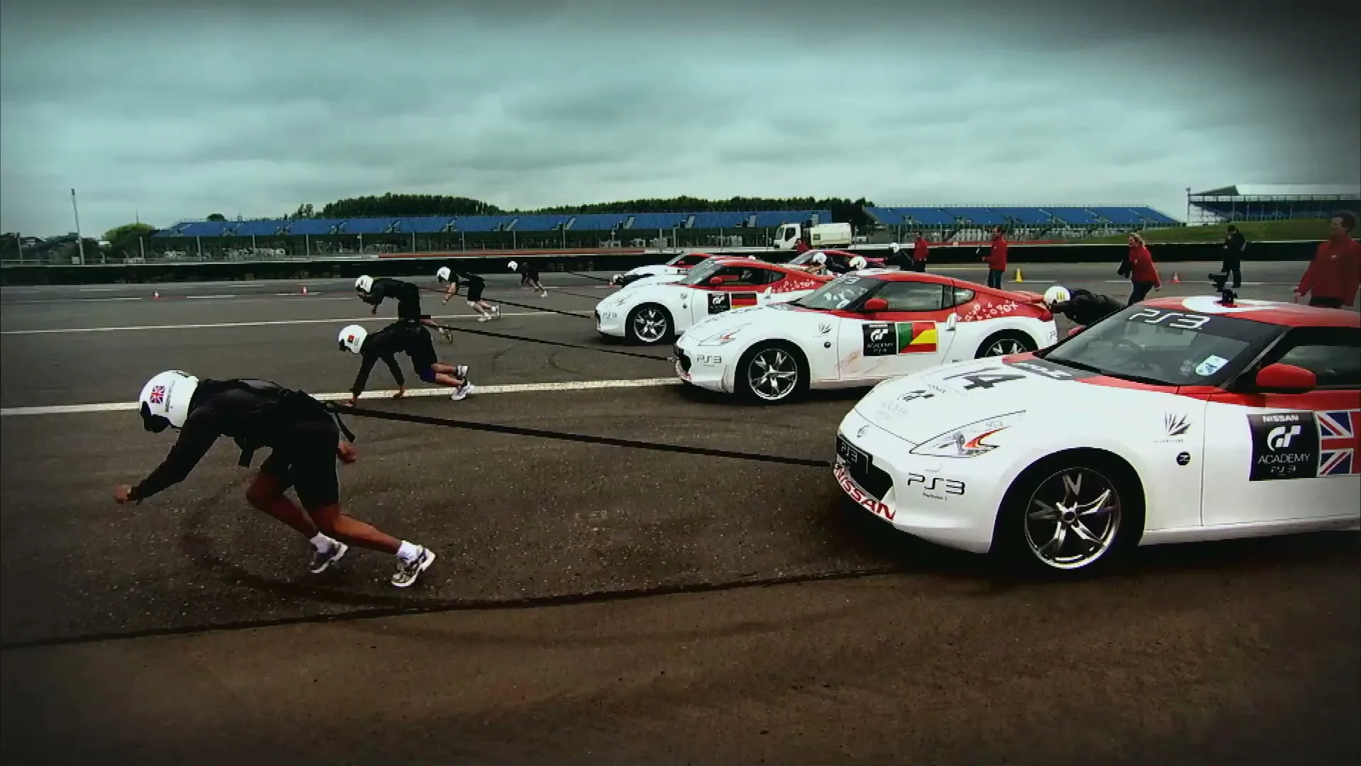 A line of male contestants in racing gear are crouched at the starting line next to Nissan sports cars on a race track under a cloudy sky.