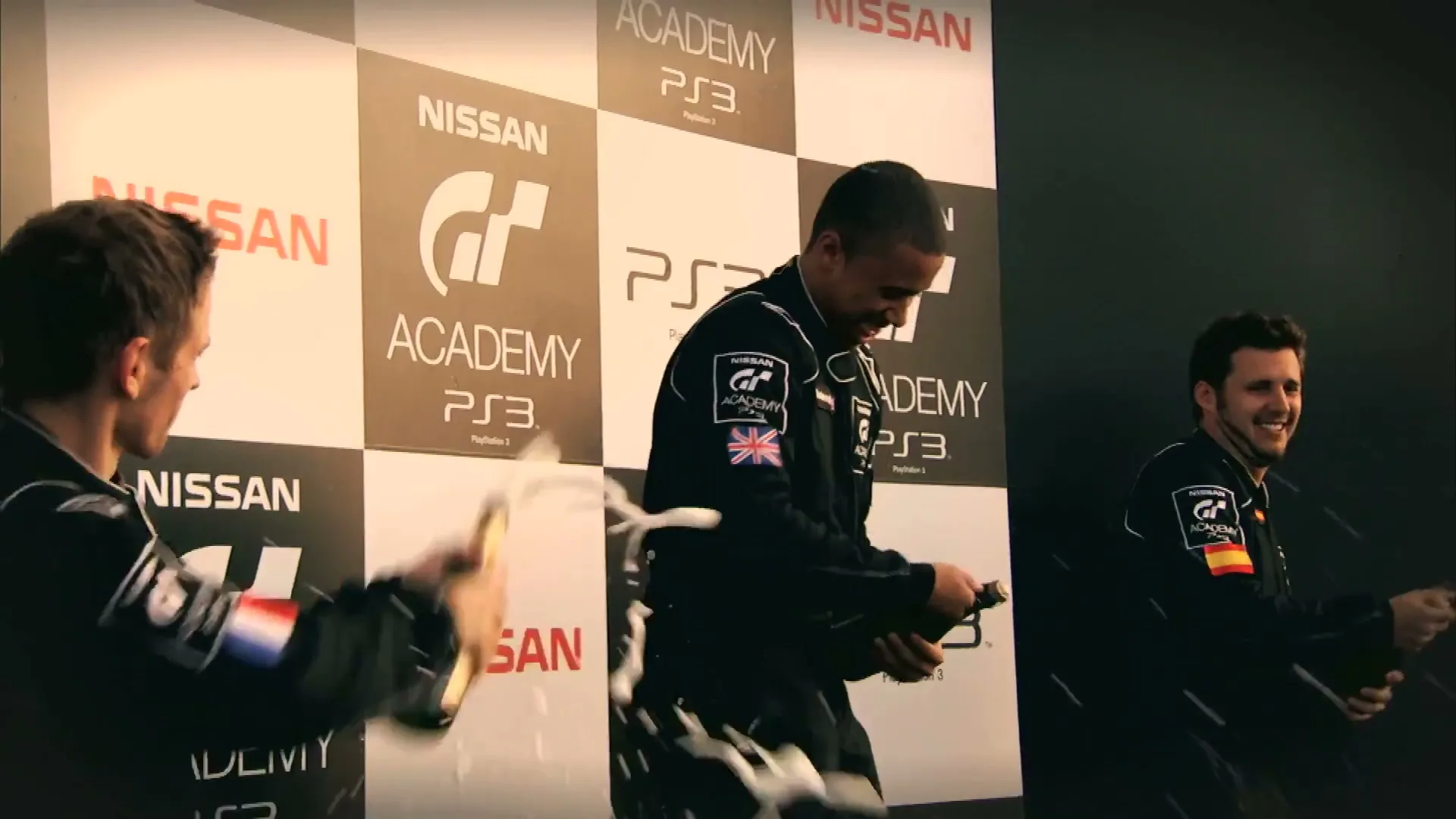 Three male race car drivers in black suits celebrate on a podium, spraying champagne, with Nissan and GT Academy logos in the background.
