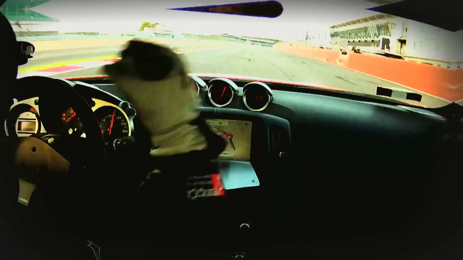 A driver's gloved hand shifts gears inside a red sports car, speeding around a race track on a bright day.