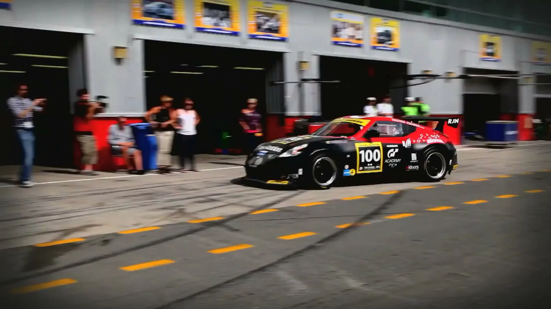 A black and red Nissan race car with "100" on the side drives out of a garage at a race track, with people watching.