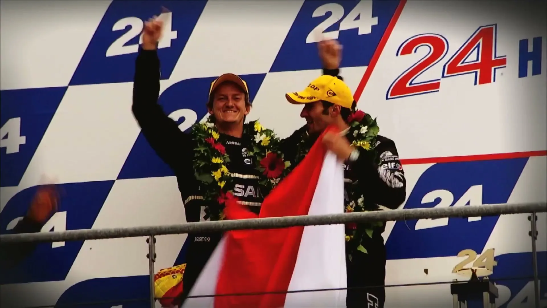 Two male race car drivers, wearing yellow caps and flower leis, celebrate on a podium with a red and white flag, against a checkered "24H" backdrop.
