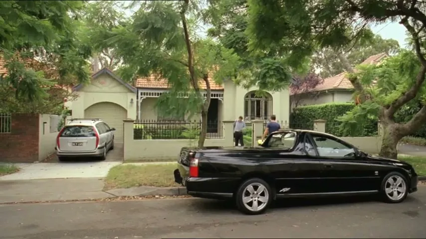 Two men stand in front of a suburban house with a black ute parked on the street and a silver station wagon in the driveway.