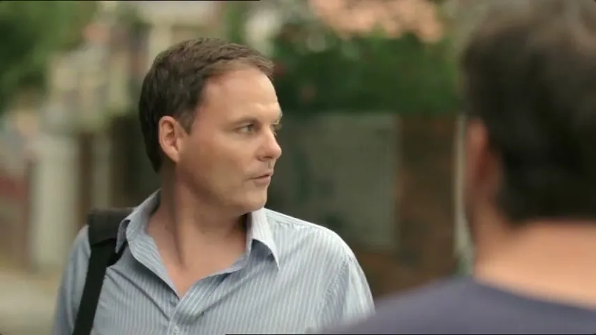 A man in a striped shirt and a black bag over his shoulder talks to another man, whose back is to the camera, on a suburban street during the day.