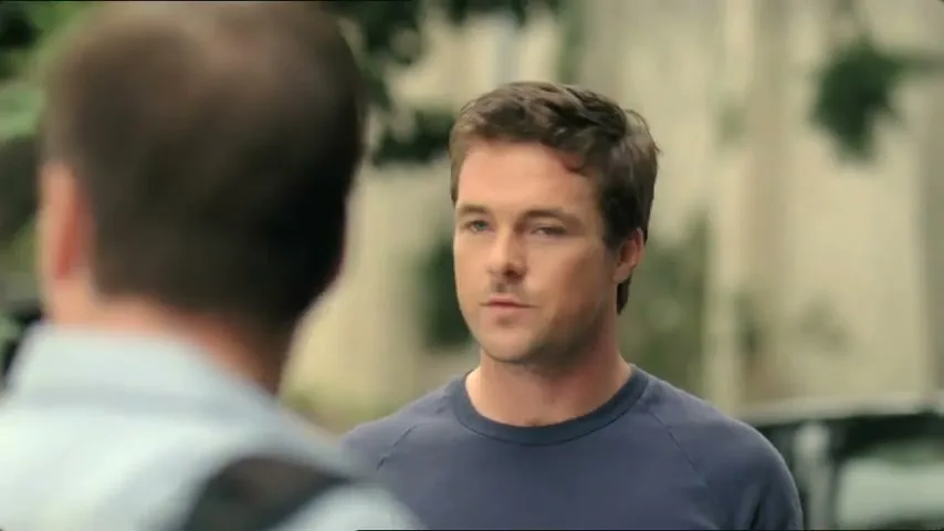 A man with brown hair and a blue shirt talks to another man, whose back is to the camera, in an outdoor setting.