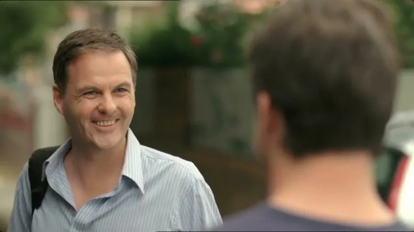 A man in a striped shirt smiles at another man, whose back is to the camera, on a suburban street.
