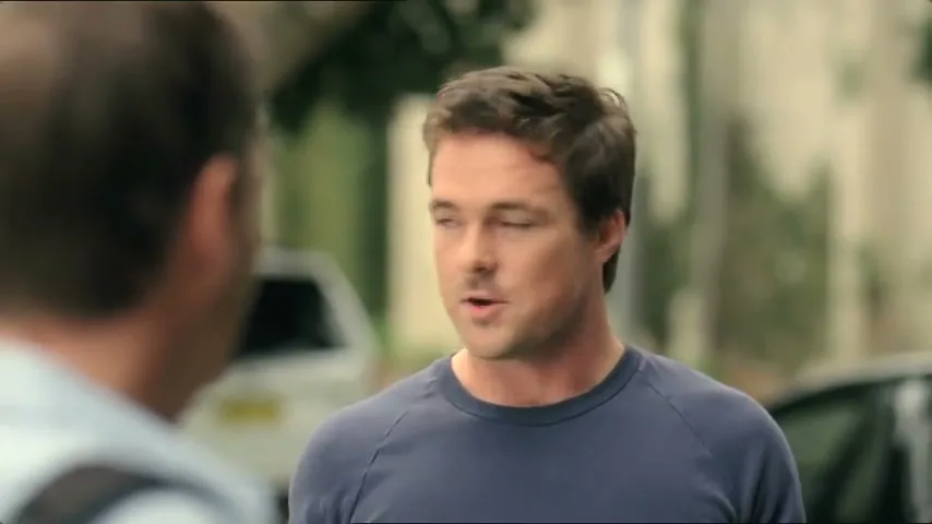 A man in a blue shirt talks to another man, then looks down, in a suburban street during the day.