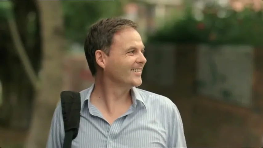 A man in a striped shirt with a bag over his shoulder smiles and looks to his right, talking to someone off-screen in an outdoor setting.