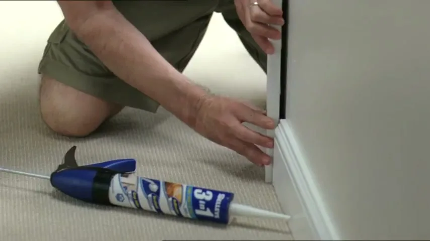 A man in khaki shorts kneels on a carpeted floor, pressing a white trim piece against a wall, with a Selleys 3-in-1 caulk gun lying nearby.