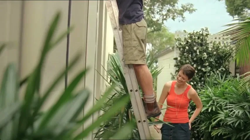 A woman in an orange top looks up at a man on a ladder, who is partially visible, in a lush green backyard.