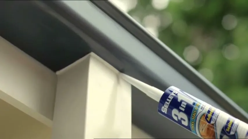 A Selleys 3 in 1 sealant tube applies white sealant to a dark grey gutter and a cream-colored wall, with green foliage blurred in the background.
