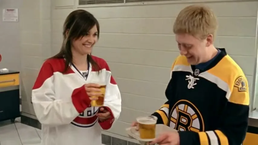 A young man in a Boston Bruins jersey offers a beer to a young woman in a Montreal Canadiens jersey in a locker room.