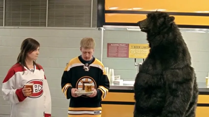 A young woman and man in hockey jerseys stand with beers, looking at a large black bear standing upright at a concession stand.