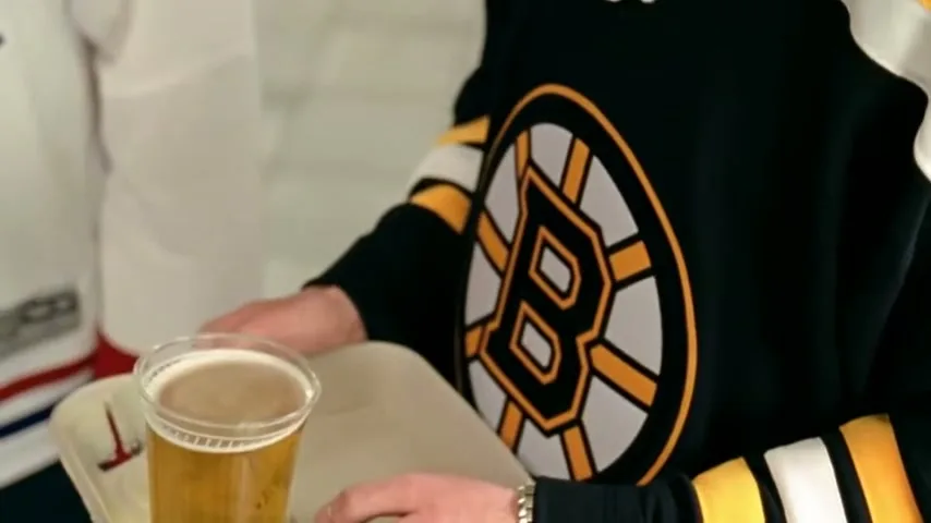 A woman in a Montreal Canadiens jersey holds a beer, then a man in a Boston Bruins jersey takes a beer from a tray, then looks at the camera in an arena concourse.