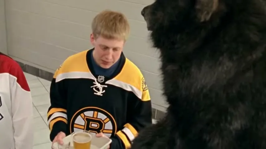 A young man in a Boston Bruins jersey holds a tray with a drink, looking at a large black bear in a locker room.