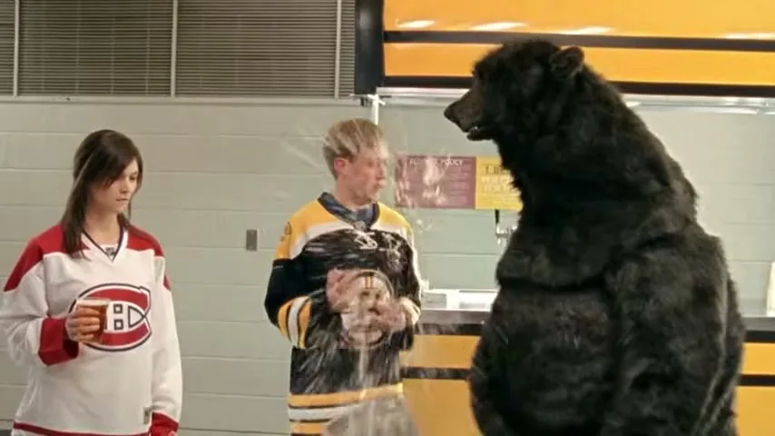 A man in a Bruins jersey gets beer spilled on him by a bear mascot, while a woman in a Canadiens jersey watches at a concession stand.