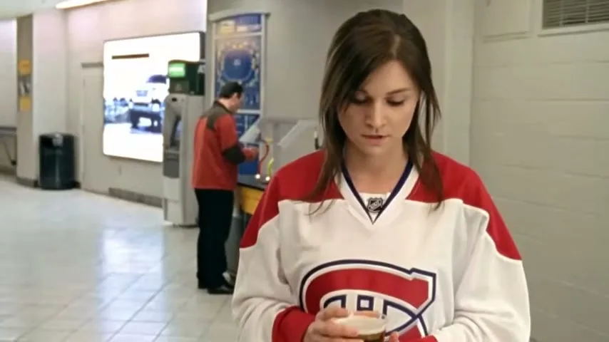A young woman in a Montreal Canadiens jersey holds a drink, looking downcast in a brightly lit arena concourse.