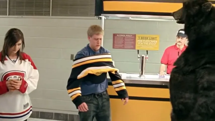 A young man in a Bruins jersey is tied up by a bear mascot, while a young woman in a Canadiens jersey watches, and a vendor stands behind a counter at a stadium.