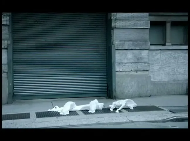 Two white, crumpled plastic bags, resembling polar bears, lie on a city sidewalk next to a building with a closed garage door, as a yellow taxi drives by.