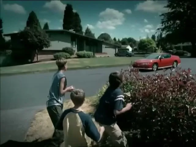 Three boys throw rocks at a red car driving down a suburban street on a sunny day.