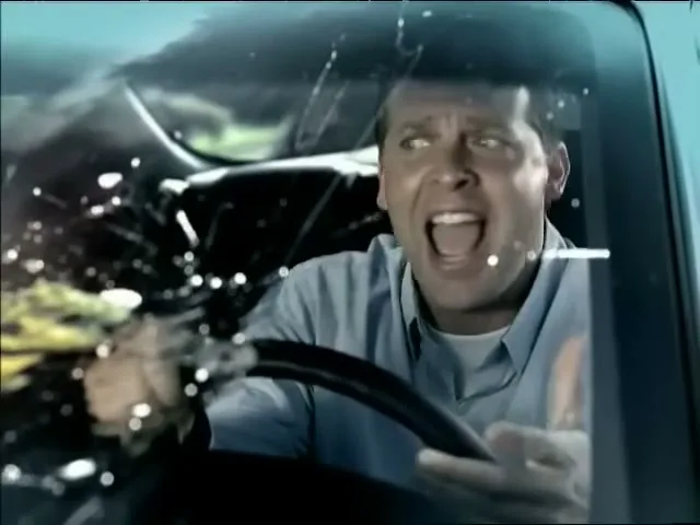 A man in his 30s, wearing a blue shirt, screams in terror as something splatters on his car windshield while he is driving.