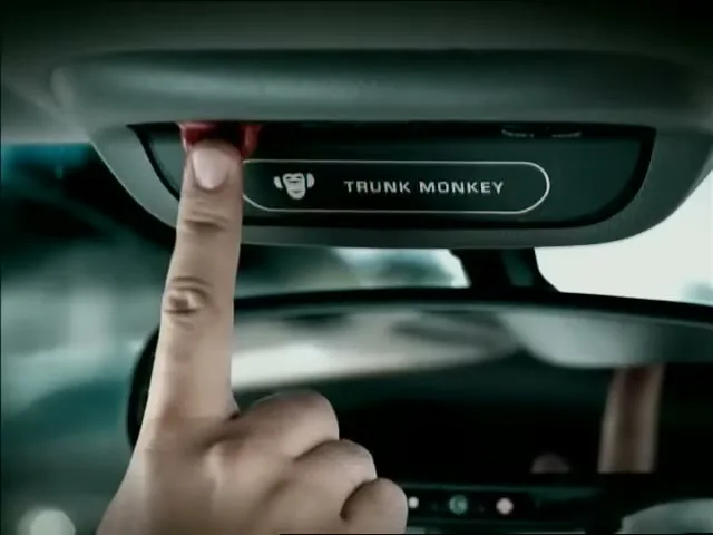 A person's finger presses a red button on a car's overhead console, activating a "Trunk Monkey" system, with the rearview mirror visible below.