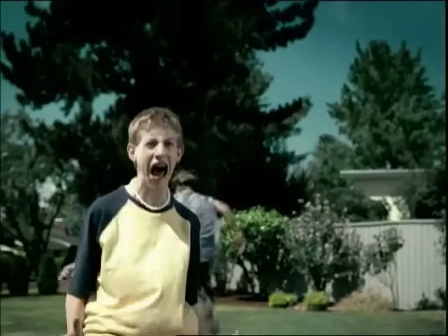 A young boy in a yellow and blue shirt screams in terror as he runs from an unseen threat in a suburban backyard.