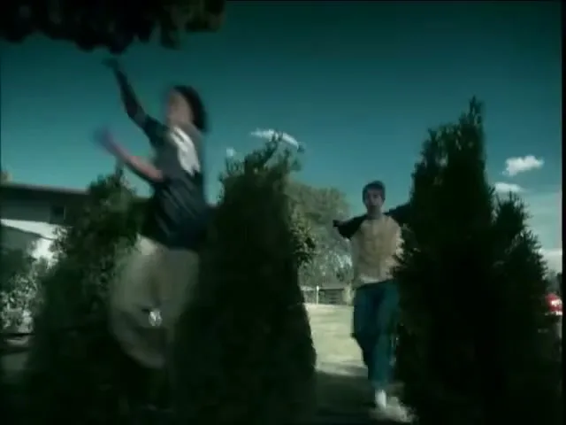 Two young men, one in a dark shirt and light pants, the other in a light shirt and dark pants, are running and jumping over bushes in a suburban yard during the day.