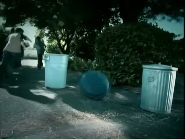 A group of people run away from a monkey in a suburban backyard, knocking over trash cans as they flee.