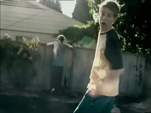A young boy in a yellow and black shirt runs towards the camera with a surprised expression, while another boy climbs a fence in the background of a suburban backyard.