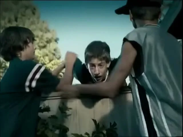 Three teen boys are seen from the chest up, one boy is being pulled over a fence by the other two boys in a suburban backyard.